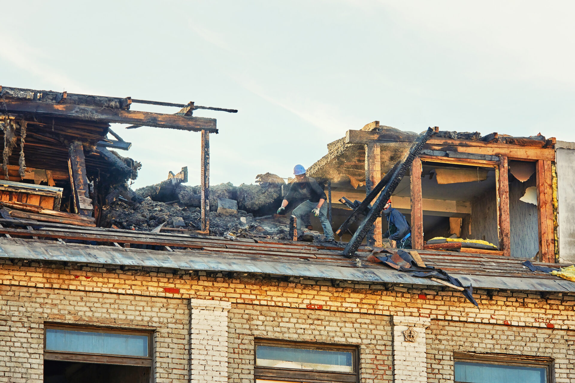 Unrecognizable construction team dismantle charred roof in the aftermath of fire. Post fire dismantling of burnt roof with team of workers. Men remove charred wooden logs from the roof